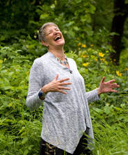 A white woman with short grey hair is holding her hands out and laughing in front of a plant-filled background. She is wearing a light grey top and necklace.