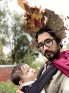 An light-skinned man with dark hair and a dark beard holds a baby to his chest. en is wearing a crown of leaves and a red cape.