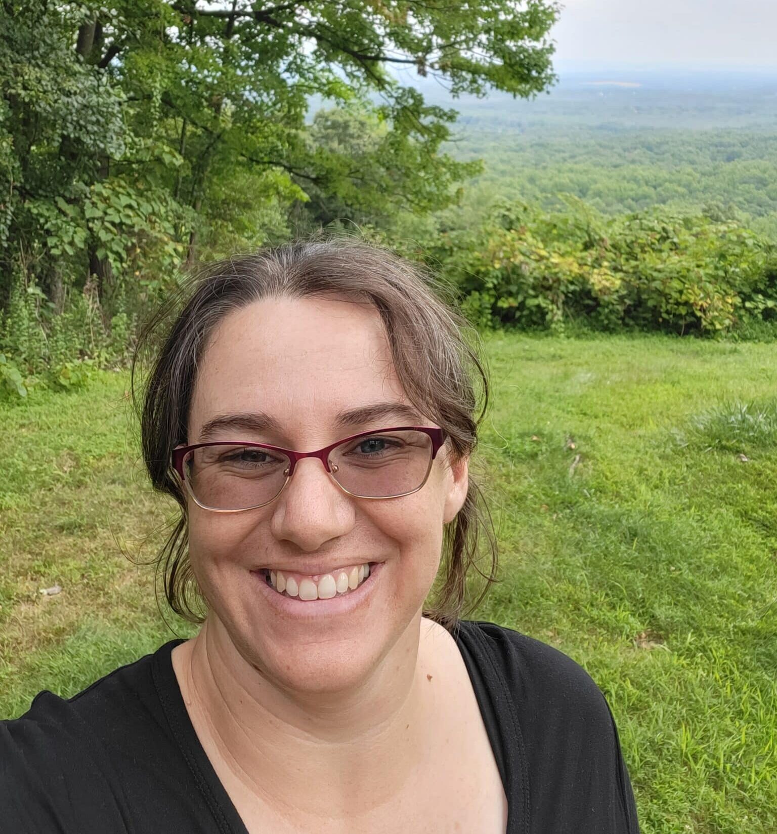 A white woman with brown hair pulled back smiles in front of a grassy background. She is wearing glasses and a black top.