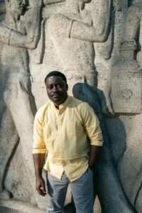 A dark-skinned man with short black hair stands in front of stone wall with carved figures on it. He is wearing a yellow button down and blue jeans.