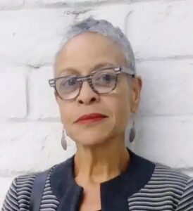 A Black woman with short grey hair, glasses, and earrings is pictured in front of a white brick wall. She is wearing a navy blue and white striped top with a navy collar.