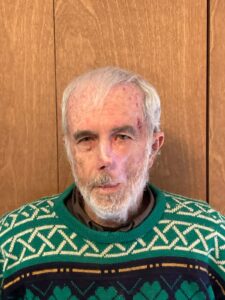 A white man with white hair and a white beard stands in front of a wooden background. He is wearing a green, black, and tan patterned sweater.
