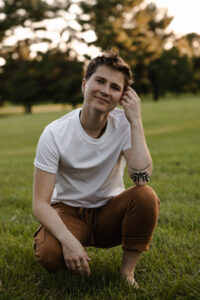 A white person in a white shirt and brown pants crouches down in a grassy field.