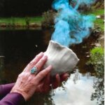 Two light-skinned hands hold a clay vessel out of which blue smoke rises.