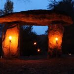 A stone megalith at dusk is light up by two torches.