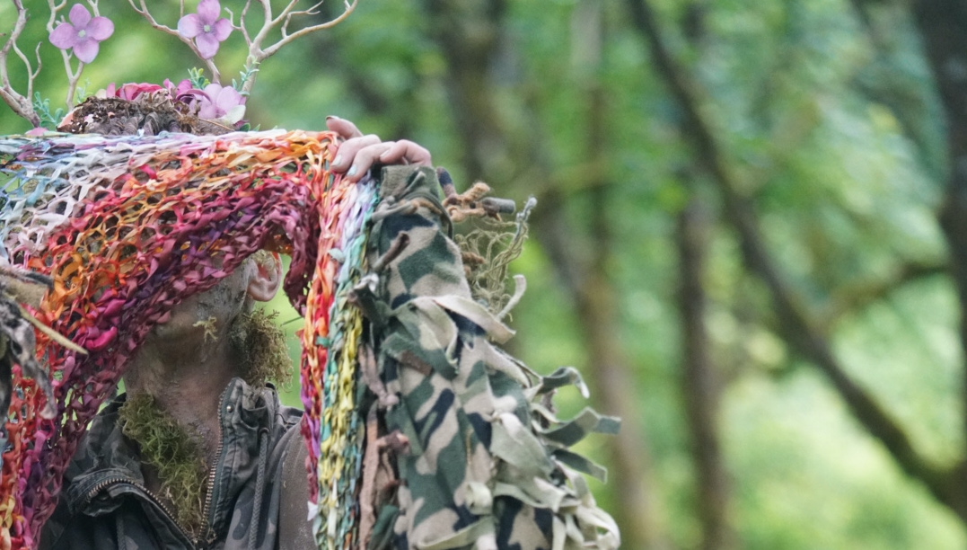 A mix of colorful and patterned fabrics hang over a tree branch in a wooded area.