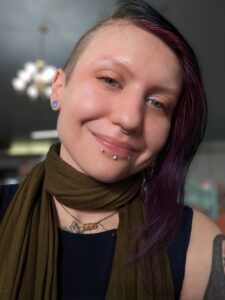A headshot of a white person with facial piercings and a purple undercut grinning at the camera.