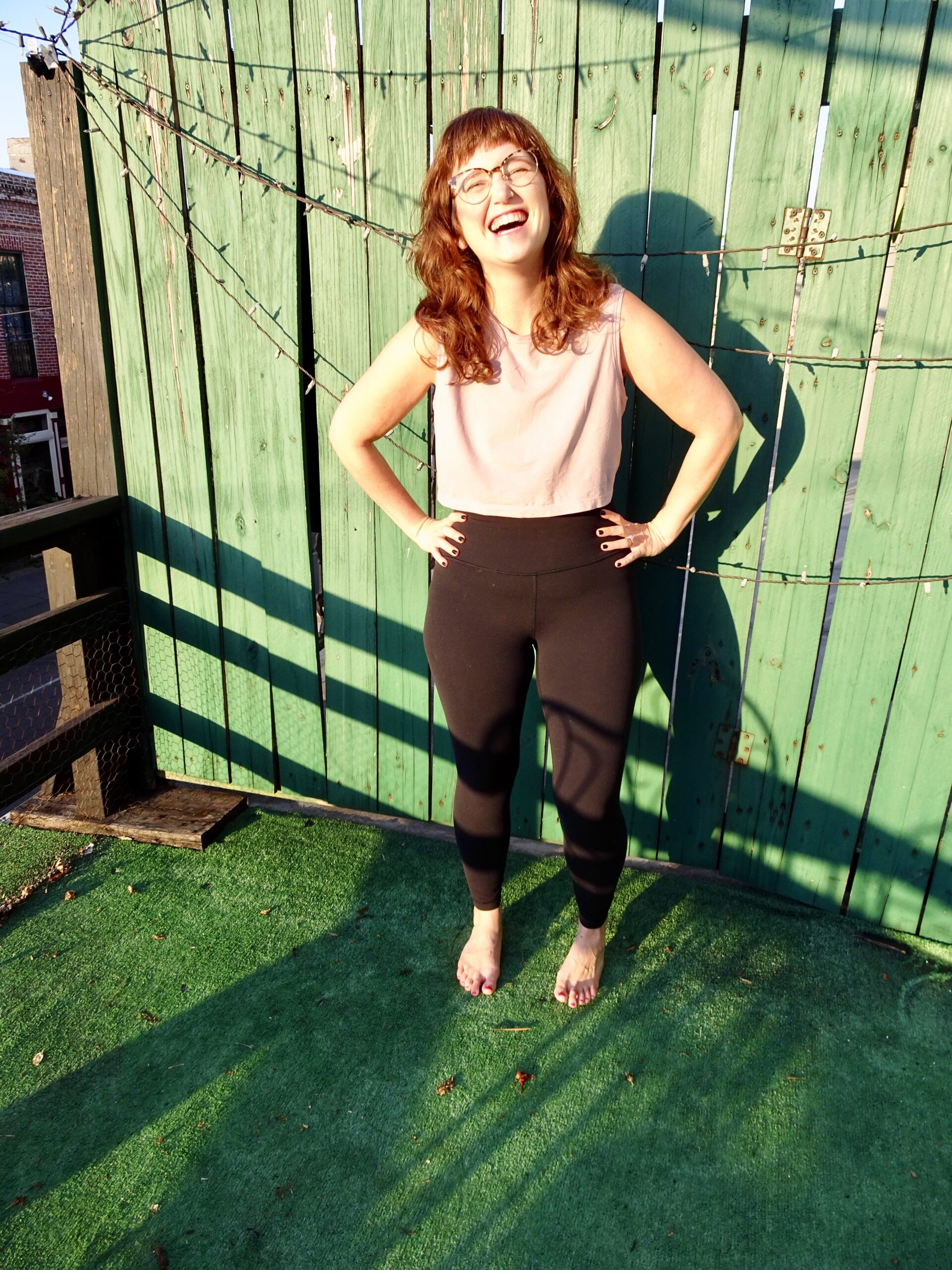White woman with long red hair laughing with her hands on her hips. Behind her is a wooden slat wall painted green.